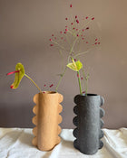 Two sculptural ceramic vases with scalloped edges, one in warm beige and one in dark charcoal gray, styled with floral stems against a taupe wall and white fabric surface.