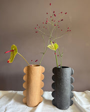 Two sculptural ceramic vases with scalloped edges, one in warm beige and one in dark charcoal gray, styled with floral stems against a taupe wall and white fabric surface.