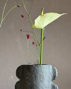 A minimalist floral arrangement featuring a pale yellow calla lily and delicate red wildflower stems in a dark textured ceramic pot against a neutral taupe background.