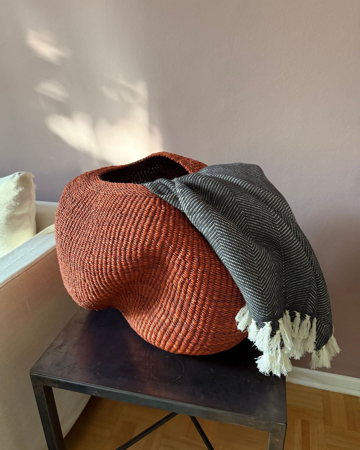 A large, organic-shaped woven basket in terracotta orange sits on a dark metal side table in a living room setting. A gray and white herringbone throw blanket with white tassels is draped over the basket.