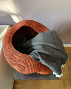 A round woven basket in a rust-orange color, styled with a gray and white herringbone patterned throw blanket, set on a dark bench in a home environment.