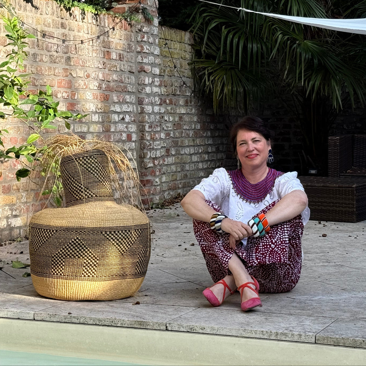 A woman sits on a stone patio beside a pool, smiling next to a large woven basket. The setting features a brick wall, potted plants, and tropical foliage in the background.