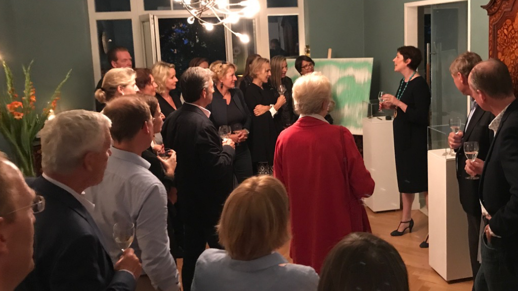 A group of people gathered indoors at what appears to be an art gallery or exhibition, standing and talking near white display pedestals. Several women in the center are facing a green canvas or print, while men in suits stand nearby holding drinks.