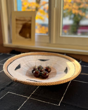 A round woven basket bowl with a natural tan rim and black geometric accents, holding a small cluster of dark red cherry tomatoes, set on a black tablecloth in front of a window.