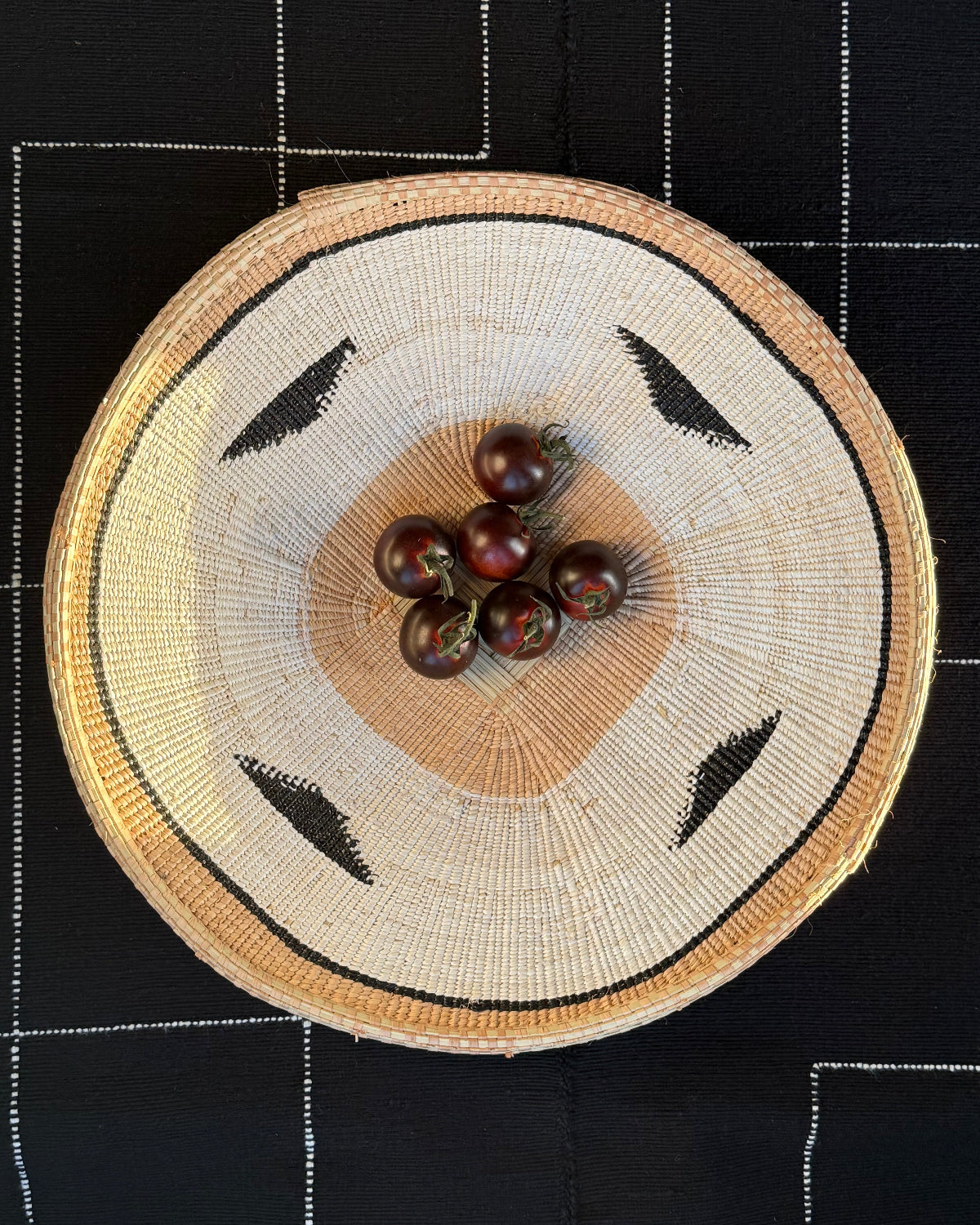 A round woven basket with a natural straw-colored body, black geometric accents, and a light tan center, photographed from above on a black fabric background with white stitched grid lines. Small dark red berries are placed in the basket.