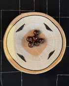 A round woven basket with a natural straw-colored body, black geometric accents, and a light tan center, photographed from above on a black fabric background with white stitched grid lines. Small dark red berries are placed in the basket.