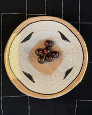 A round woven basket with a natural straw-colored body, black geometric accents, and a light tan center, photographed from above on a black fabric background with white stitched grid lines. Small dark red berries are placed in the basket.