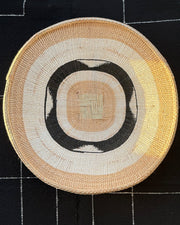 A round woven basket with concentric patterns in beige, cream, black, and yellow, displayed on a black fabric background with white stitched grid lines.