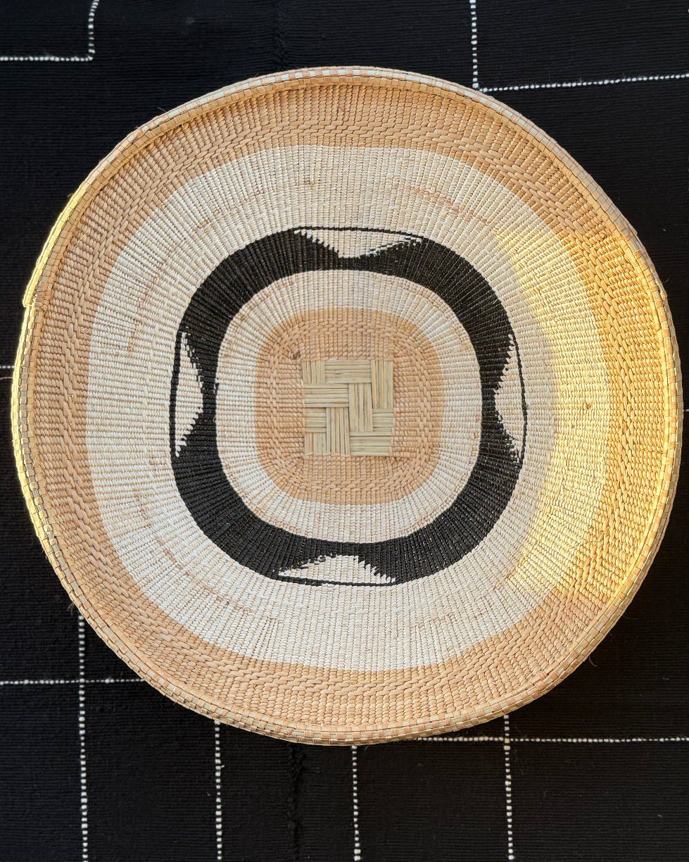 A round woven basket with concentric patterns in beige, cream, black, and yellow, displayed on a black fabric background with white stitched grid lines.