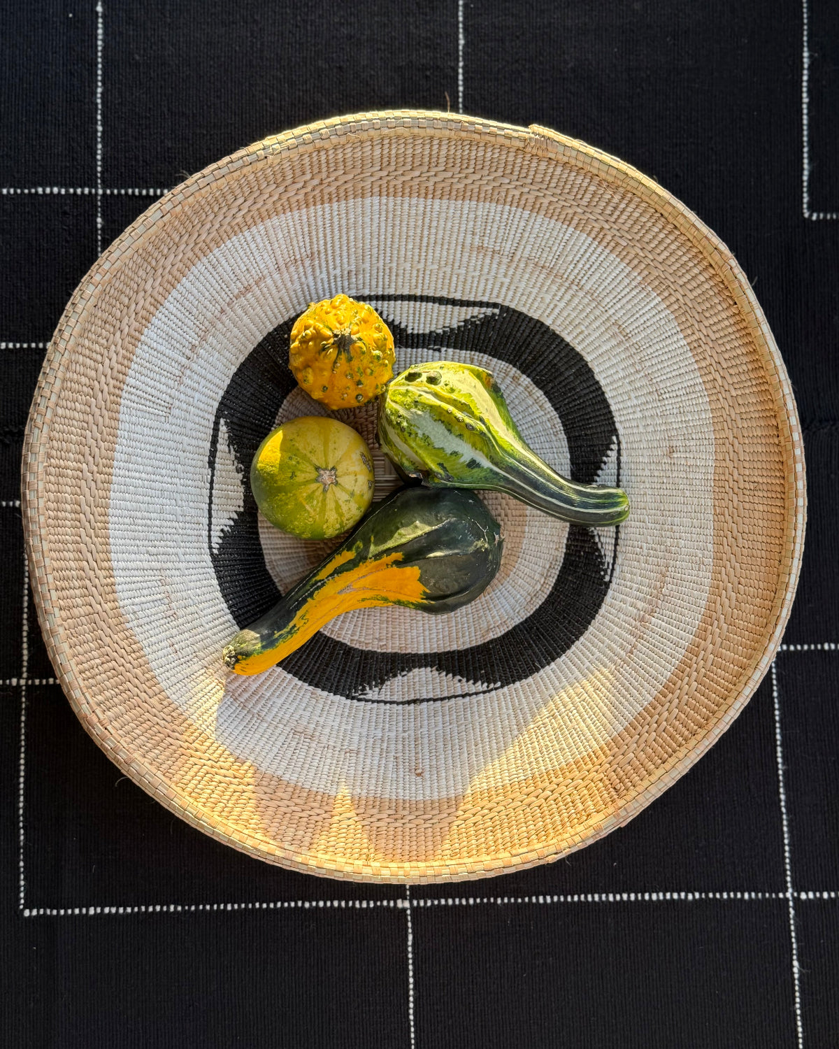 A round woven straw plate with a black and white circular pattern, styled with four decorative gourds on top, set against a black tablecloth with white grid lines.