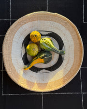 A round woven straw plate with a black and white circular pattern, styled with four decorative gourds on top, set against a black tablecloth with white grid lines.