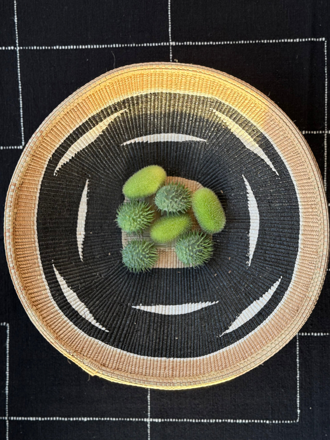 A top-down view of a round woven basket or tray with a black and white geometric pattern and a natural tan rim, holding five small green spiky fruits on a black checkered fabric background.