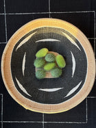 A top-down view of a round woven basket or tray with a black and white geometric pattern and a natural tan rim, holding five small green spiky fruits on a black checkered fabric background.