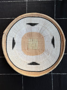 A round hand-woven basket with a geometric pattern, featuring a beige center, white outer ring, and black accents, displayed on a black and white patterned fabric background.