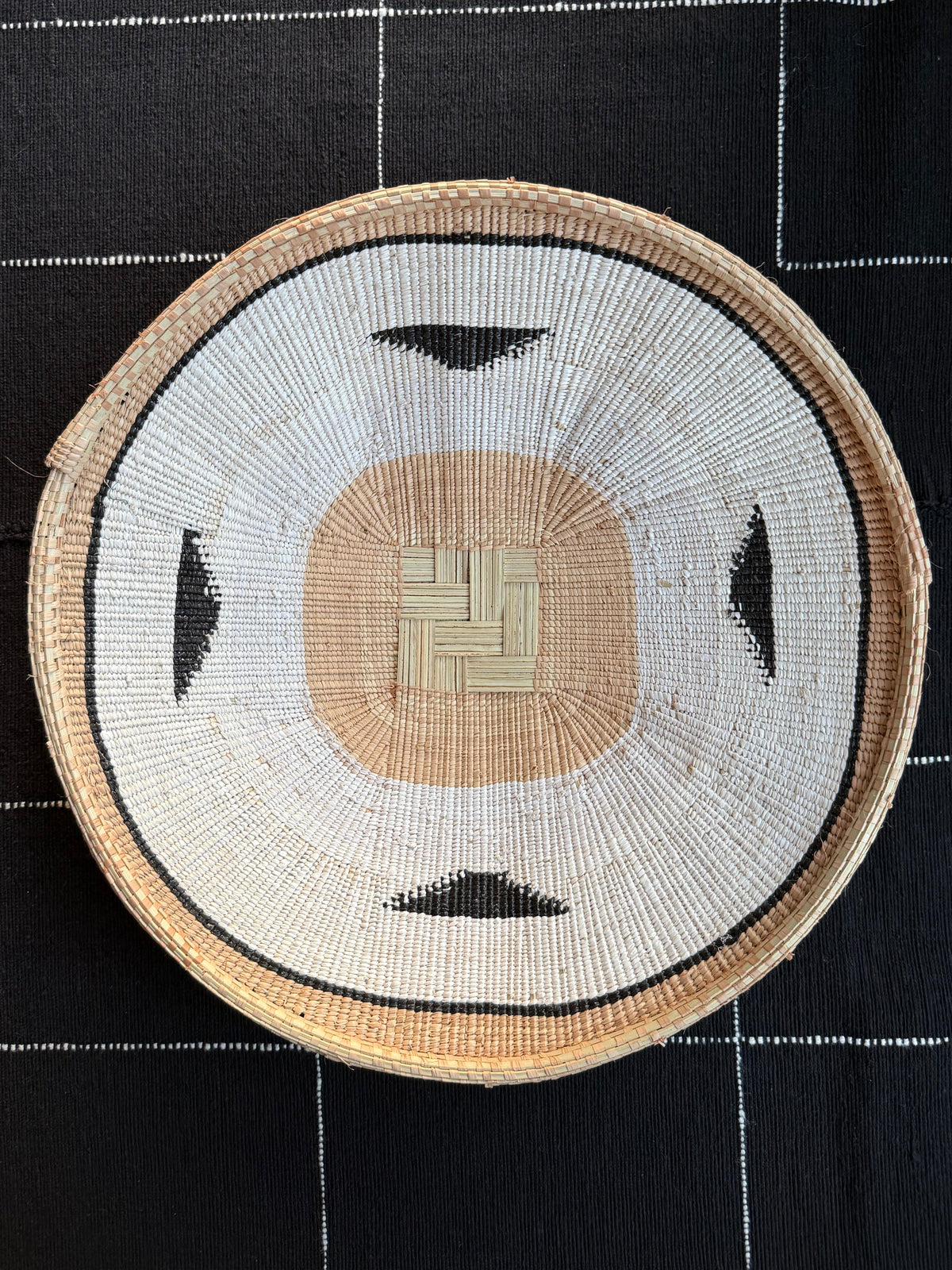 A round hand-woven basket with a geometric pattern, featuring a beige center, white outer ring, and black accents, displayed on a black and white patterned fabric background.