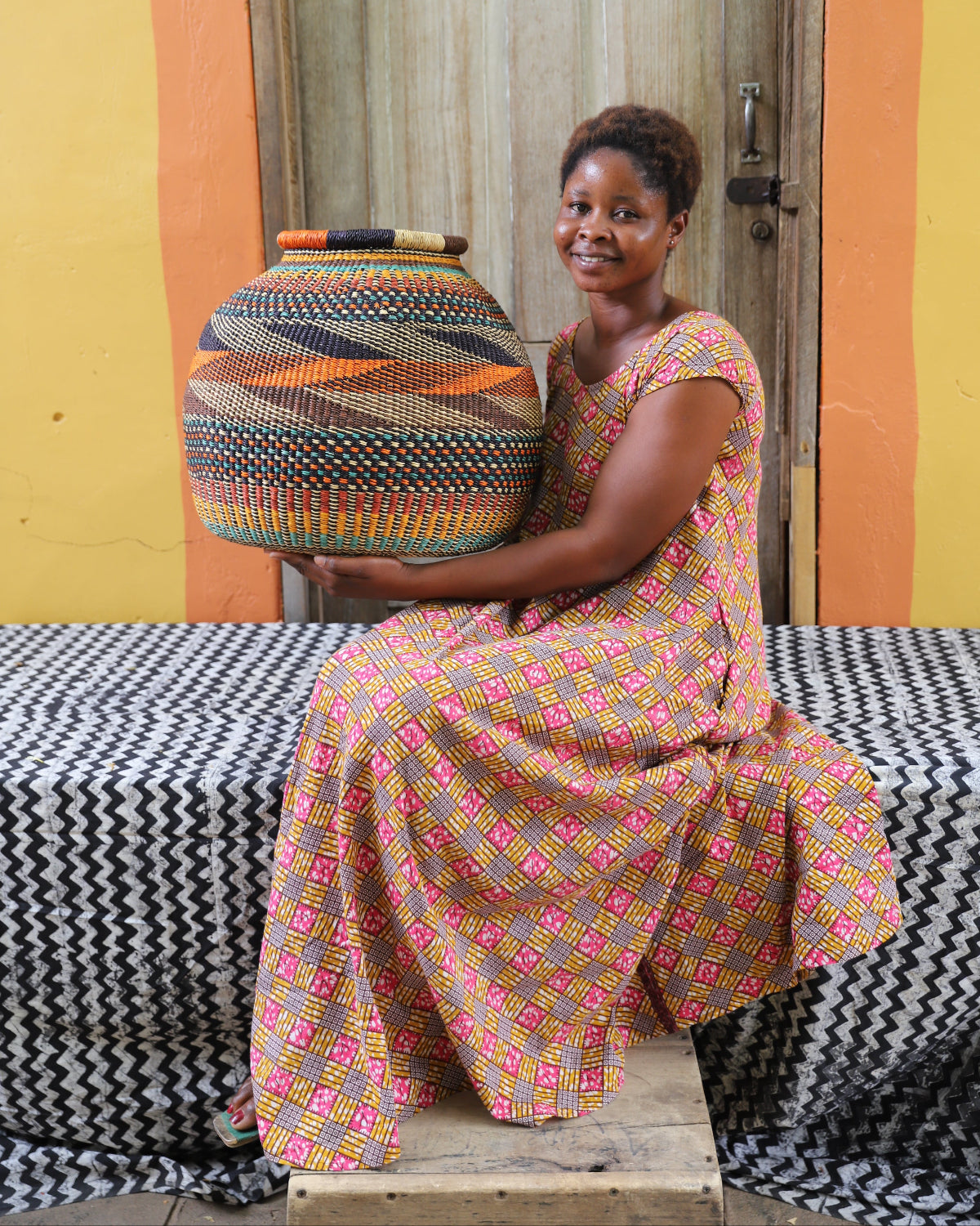 An artisan sitting with a colorful patterned basket on their lap, against a backdrop of a colorful wall and a wooden door.