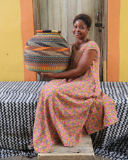 An artisan sitting with a colorful patterned basket on their lap, against a backdrop of a colorful wall and a wooden door.