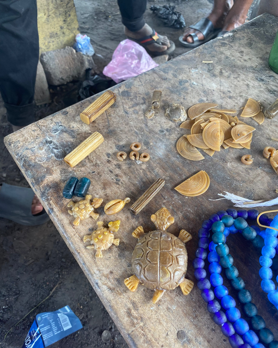 A rustic wooden table displays various handmade carvings and jewelry, including a carved turtle, small yellow stone rings, blue and purple beaded strands, and carved rectangular pieces, with parts of people's legs and feet visible around the table.