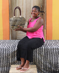 A woman wearing a bright pink tank top and black pants holds a woven straw handbag with a dark handle, posed in front of a colorful yellow and orange wall and a wooden door.