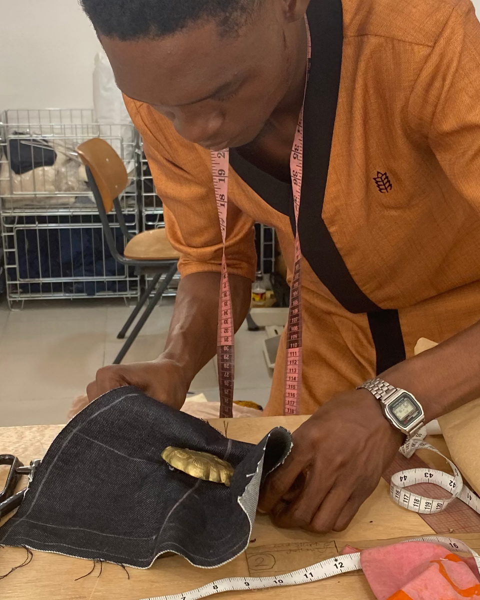 Tailor working at a table, adjusting a dark denim garment with a gold clasp. 