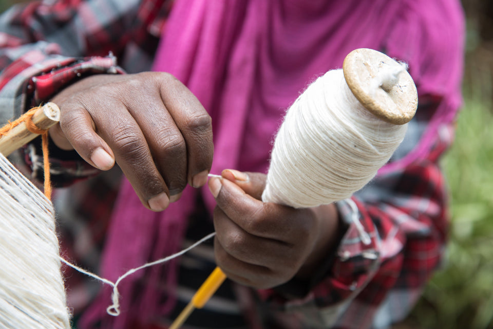 A close-up of a person's hands spinning white thread on a wooden spindle, wearing a pink and red plaid shirt.