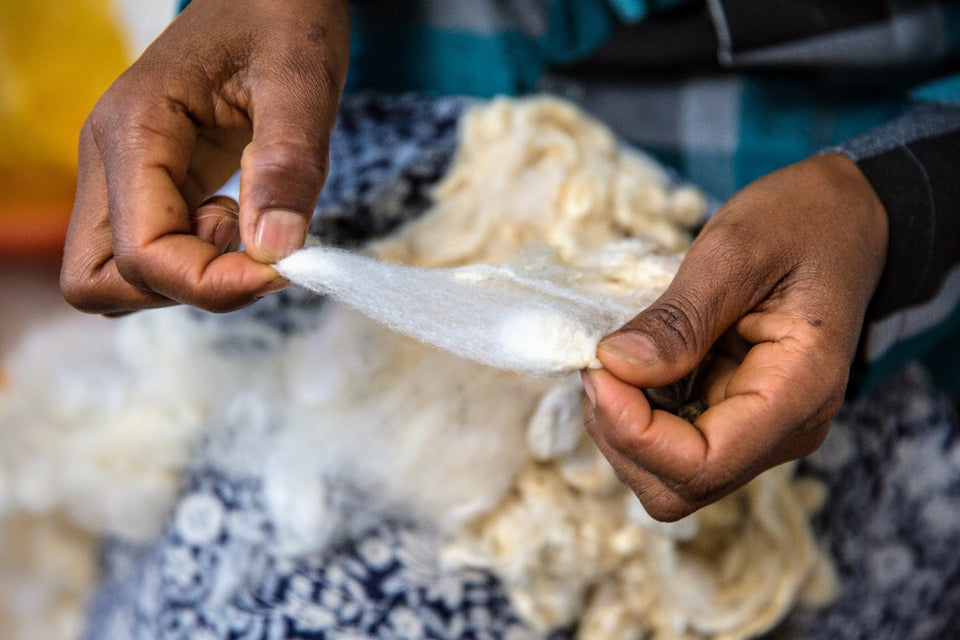Close-up of a person's hands holding and stretching a piece of white cotton or wool fabric, with more loose white fibers visible in the background.