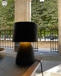 A modern table lamp with a black paper pulp drum shade and a textured dark base, illuminated with a warm yellow glow, set on a wooden table in a covered outdoor walkway.
