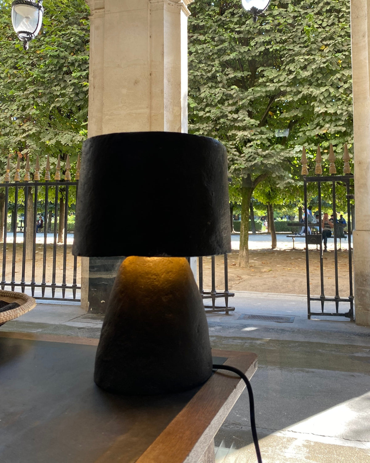 A modern table lamp with a black paper pulp drum shade and a textured dark base, illuminated with a warm yellow glow, set on a wooden table in a covered outdoor walkway.