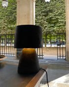 A modern table lamp with a black paper pulp drum shade and a textured dark base, illuminated with a warm yellow glow, set on a wooden table in a covered outdoor walkway.