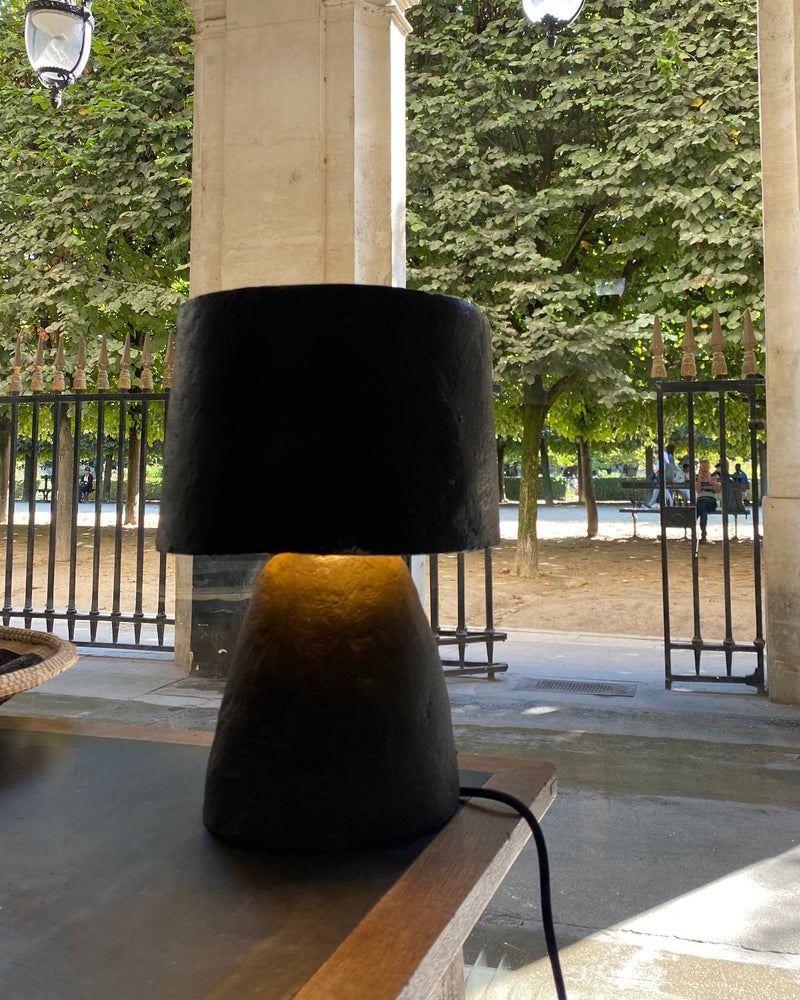 A modern table lamp with a black paper pulp drum shade and a textured dark base, illuminated with a warm yellow glow, set on a wooden table in a covered outdoor walkway.