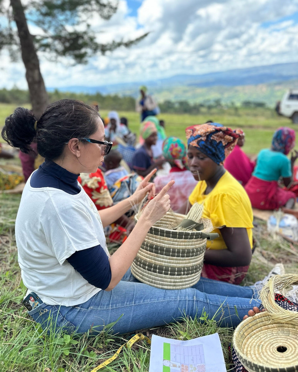 A woman in a white and navy t-shirt sits on the grass outdoors, weaving a round woven basket with straw and dark patterned strips. Other people and woven baskets are visible in a grassy outdoor setting under a cloudy sky.