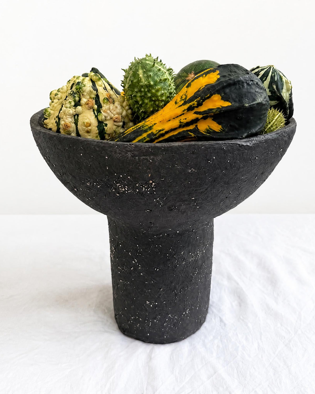 A dark charcoal gray paper pulp pedestal bowl filled with assorted decorative gourds and squash, set against a plain white background.
