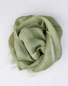 A light sage green scarf, draped and folded into a loose rosette shape, displayed on a clean white textured fabric background.