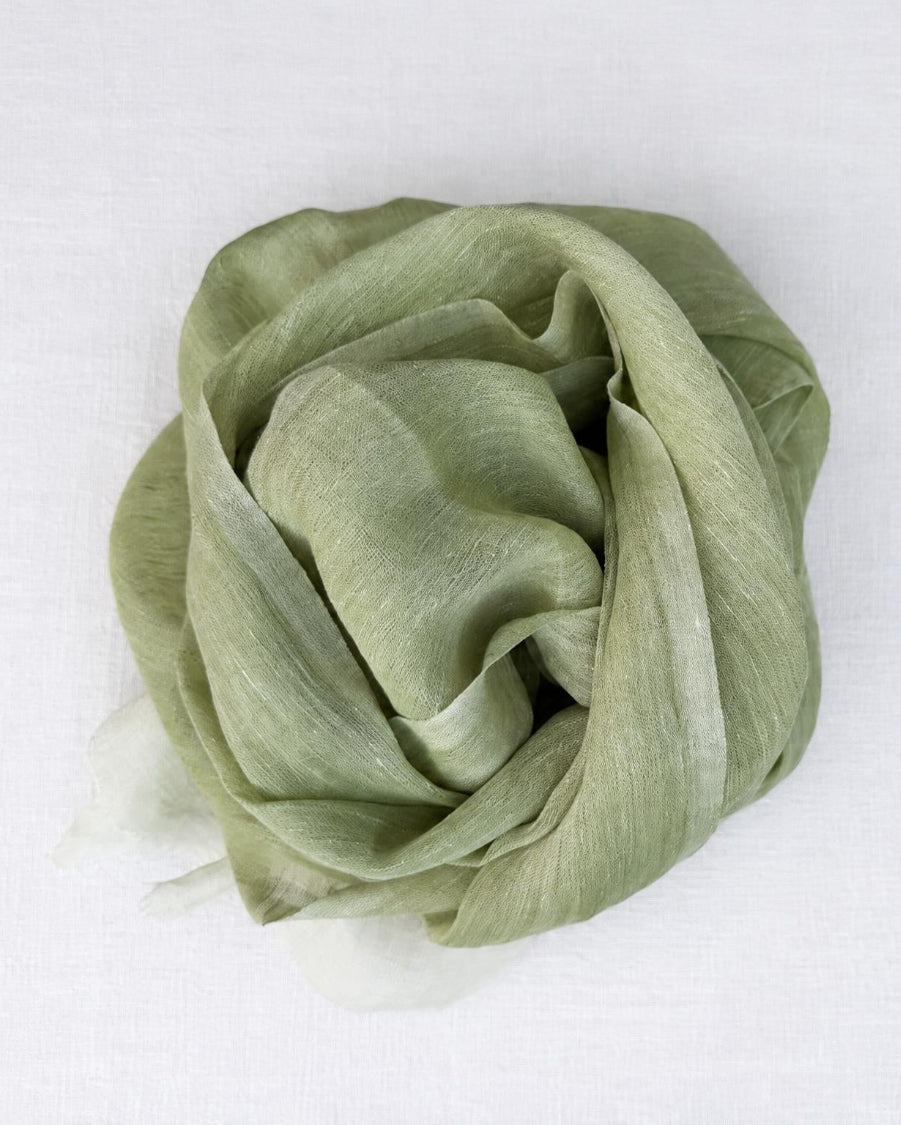 A light sage green scarf, draped and folded into a loose rosette shape, displayed on a clean white textured fabric background.
