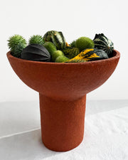 A terracotta-colored paper pulp pedestal bowl filled with decorative gourds and greenery, photographed against a clean white studio background.