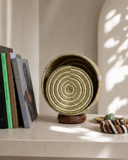 A woven round lamp with a spiral pattern sits on a wooden stand on a shelf, styled with books and a small striped accessory under soft shadow light.