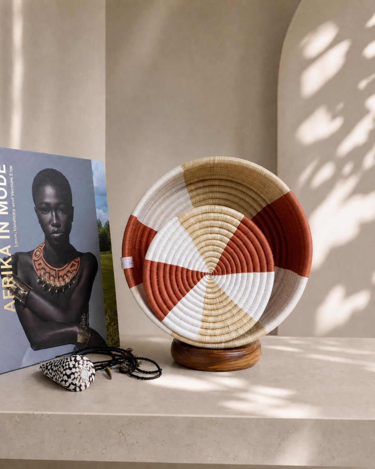 A woven decorative basket with alternating beige, terracotta red, and white segments sits on a wooden stand on a stone surface. A book titled 'AFRIKA IN MODE' and a small patterned accessory are placed beside it in a warm, sunlit interior setting with soft shadows.
