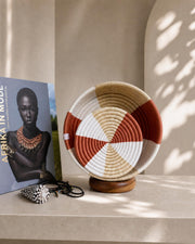 A woven decorative basket with alternating beige, terracotta red, and white segments sits on a wooden stand on a stone surface. A book titled 'AFRIKA IN MODE' and a small patterned accessory are placed beside it in a warm, sunlit interior setting with soft shadows.