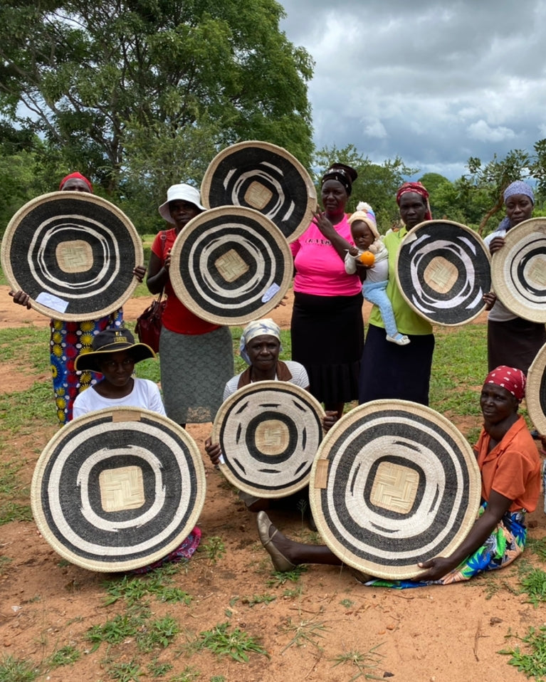 A group of people outdoors holding round woven baskets with black, white, and beige concentric patterns. Trees, grass, and a cloudy sky are visible in the background.