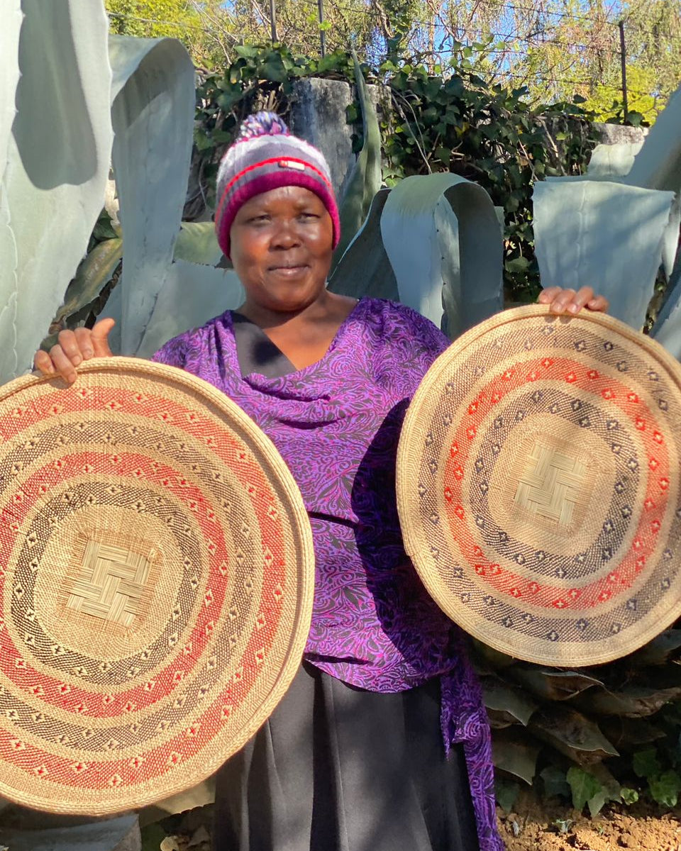 A woman wearing a purple patterned top and a pink and white knit hat holds up two round woven baskets with concentric circular patterns in beige, red, and brown, set against a background of large green leaves.