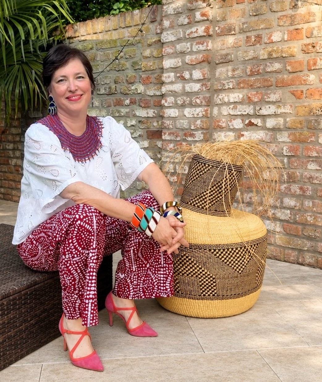 A woman sits on a dark woven outdoor sofa next to a large round woven pouf in a patio setting with a brick wall and palm leaves. She is wearing a white short-sleeve blouse, red-and-white patterned pants, and pink high heels.
