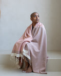 A woman sits barefoot on a minimalist beige stone step, wrapped in a dusty pink woven throw blanket with cream fringe and striped borders.