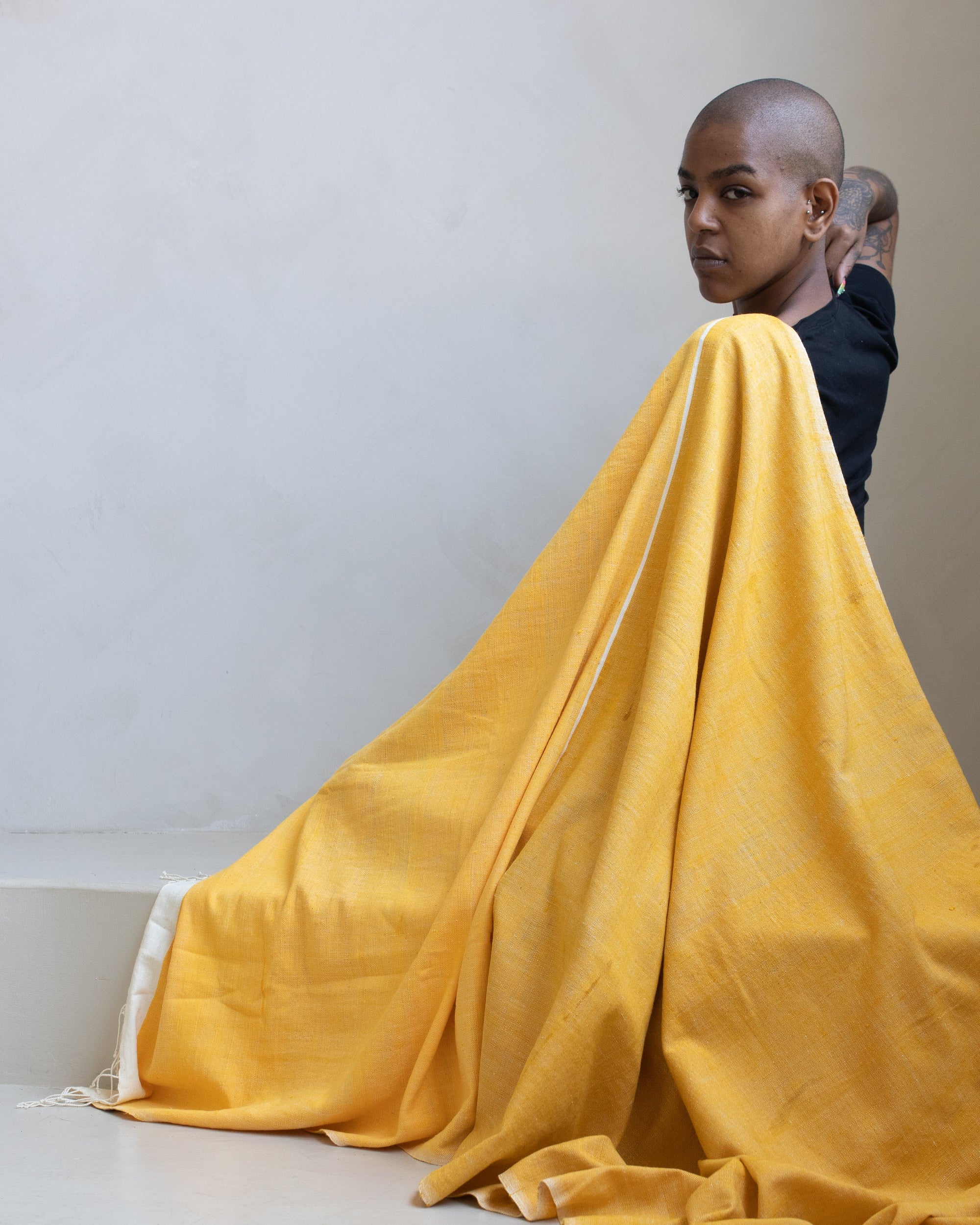 A person draped in a large yellow handwoven towel, shown from behind and side, with the fabric cascading over their shoulders and pooling on the floor against a plain light beige wall.