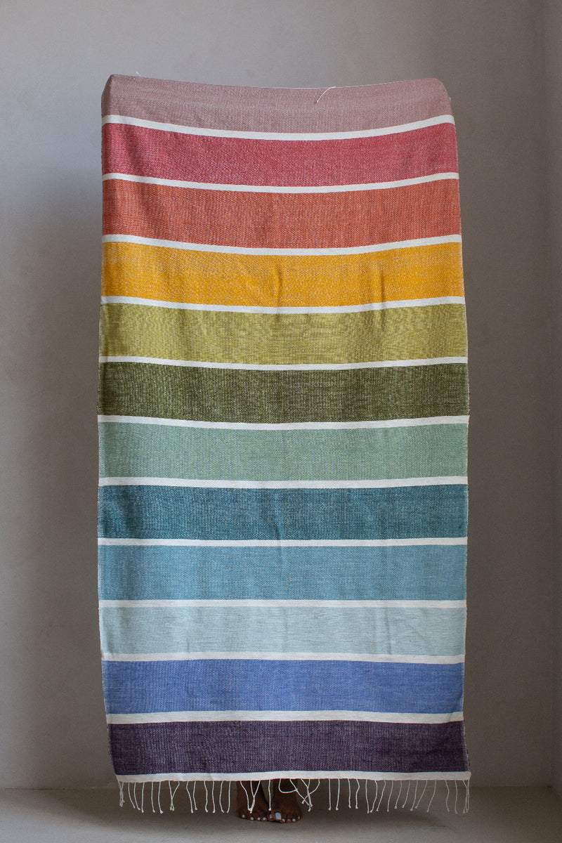 A rectangular woven textile with horizontal rainbow stripes in pink, orange, yellow, olive green, teal, blue, and purple, hanging against a plain gray wall with fringe along the bottom edge.