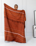 A person standing in a room with a white background, holding up a copper striped throw blanket with fringes.