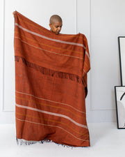 A person standing in a room with a white background, holding up a copper striped throw blanket with fringes.