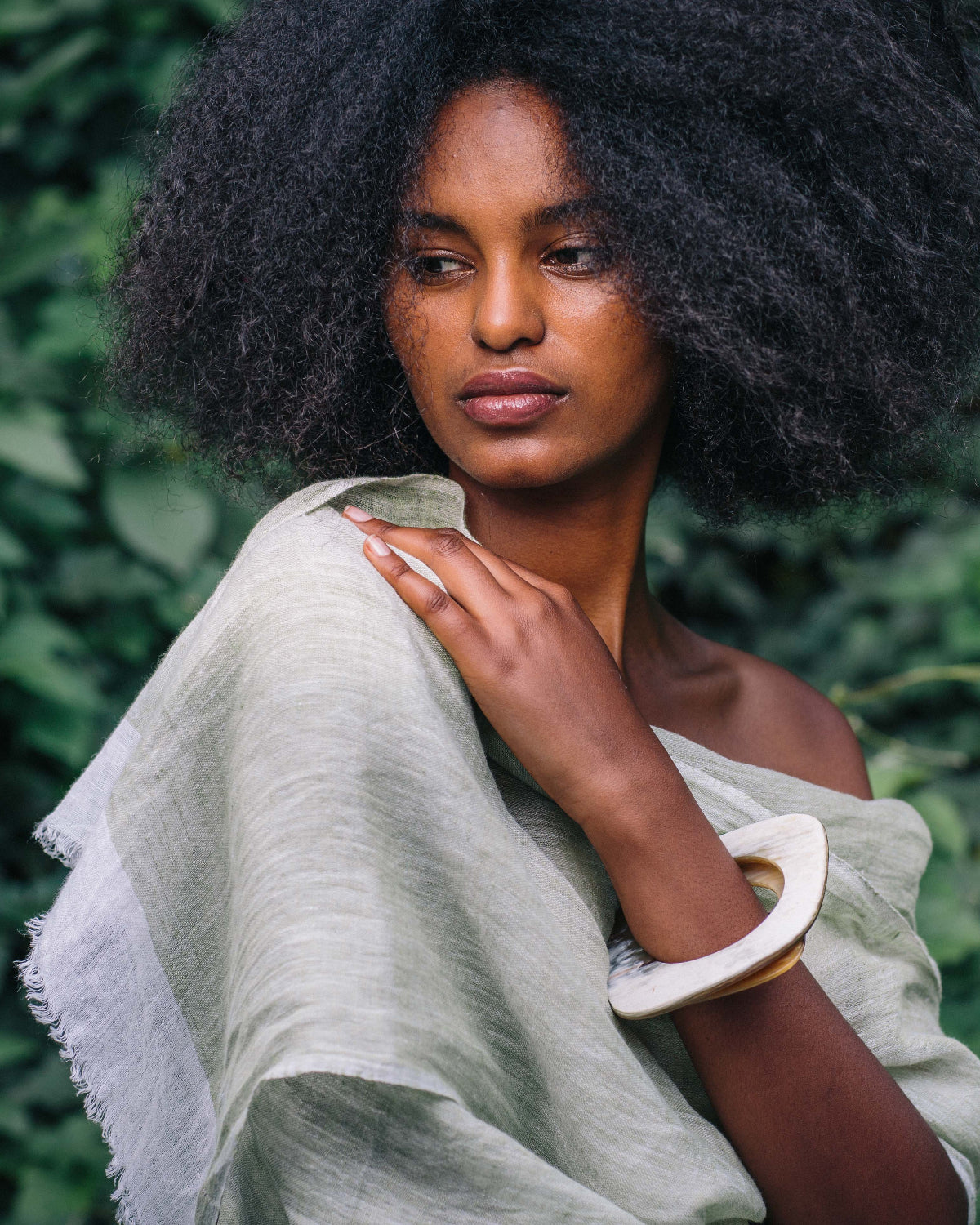 A woman with a large dark afro hairstyle is shown outdoors against a background of green foliage. She is wearing a light sage green draped shawl or wrap and a thick white bangle.