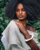 A woman with a large dark afro hairstyle is shown outdoors against a background of green foliage. She is wearing a light sage green draped shawl or wrap and a thick white bangle.