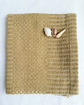 A folded Bronze-coloured woven hand towel displayed on a white background with three small seashells placed on the upper right corner.