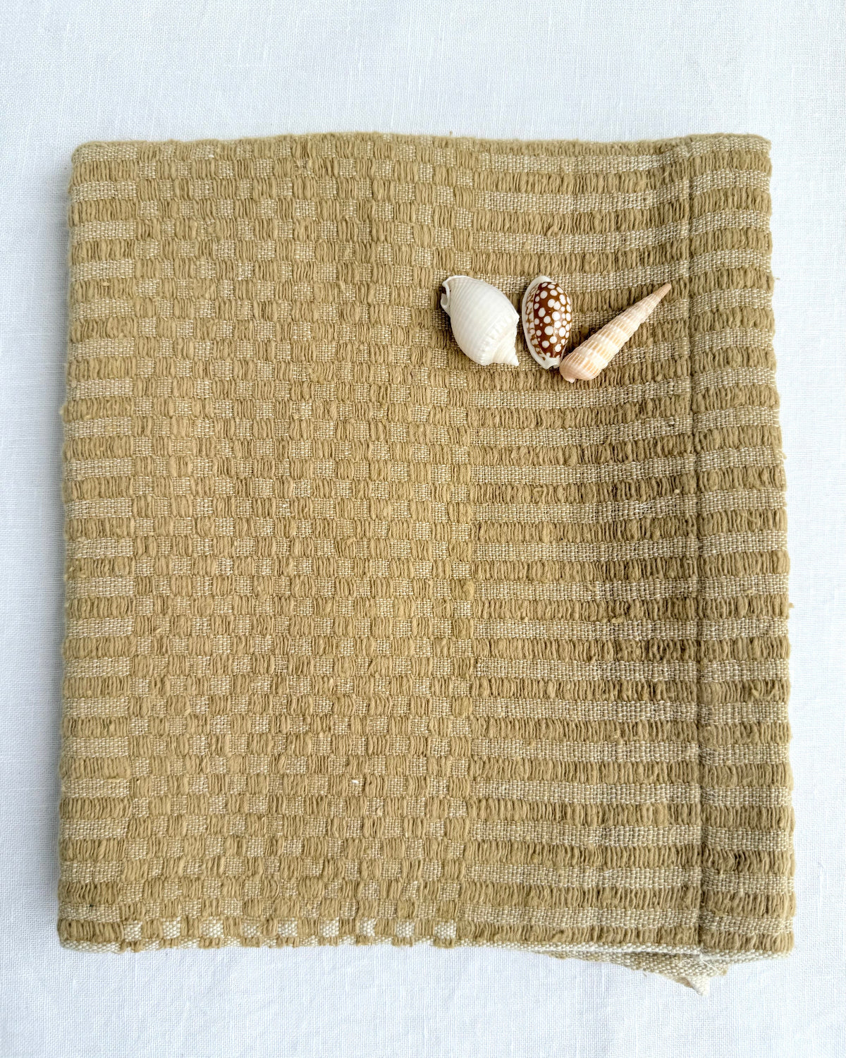 A folded Bronze-coloured woven hand towel displayed on a white background with three small seashells placed on the upper right corner.